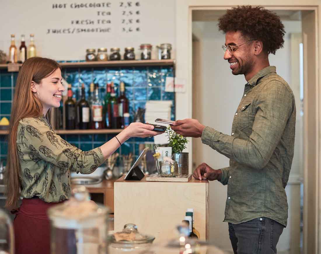 Ein Mann bezahlt in einem Coffee Shop lächelnd mit seinem Handy bei einer freundlichen Kassiererin