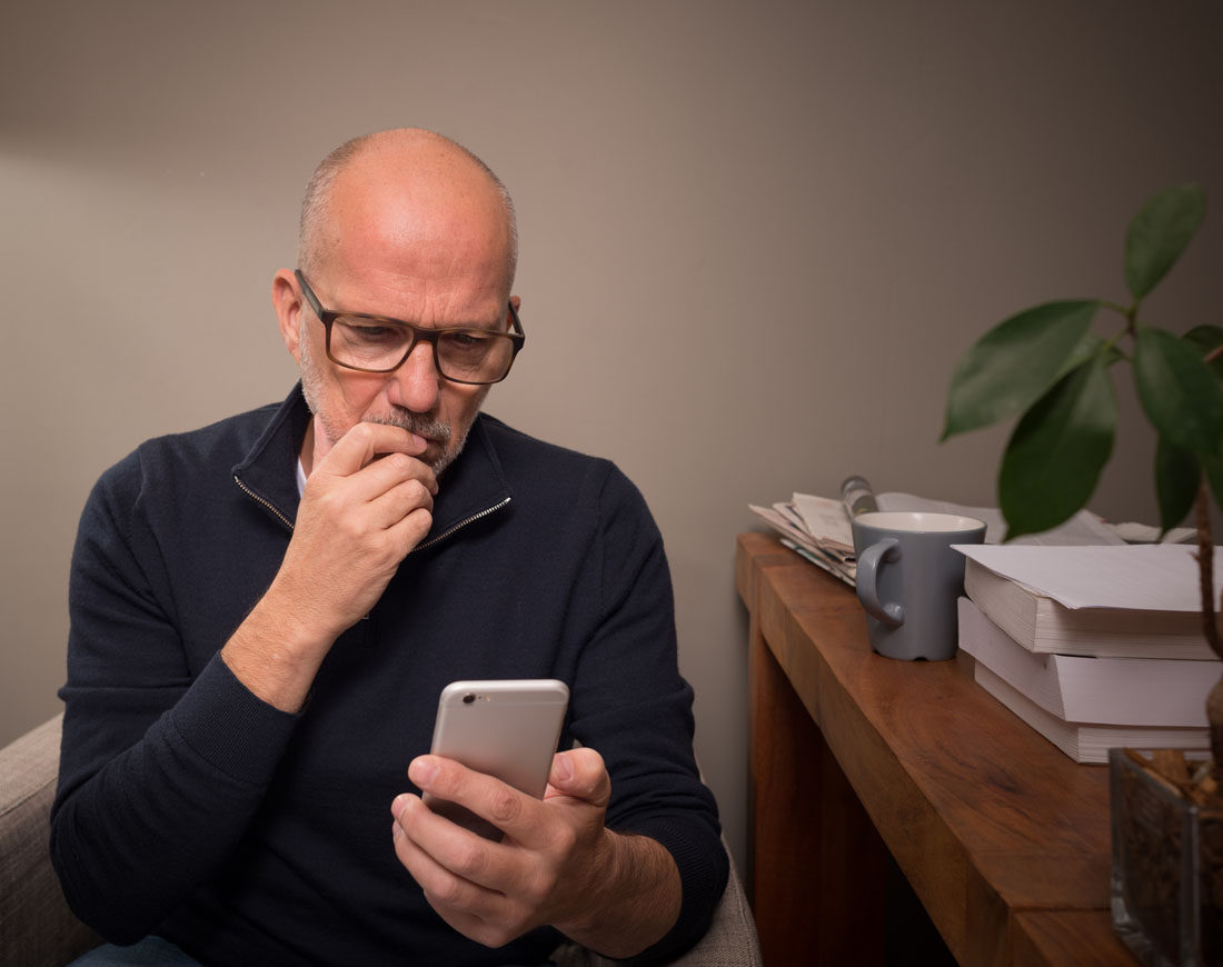 Ein Mann mittleren Alters sitzt zu Hause auf einem Stuhl und blickt kritisch auf sein Smartphone