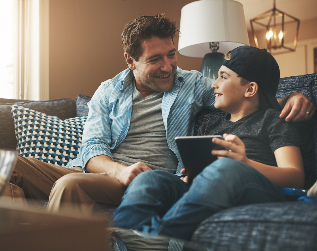 Vater und Sohn sitzen mit Tablet auf einem Sofa und unterhalten sich über Bank-Apps