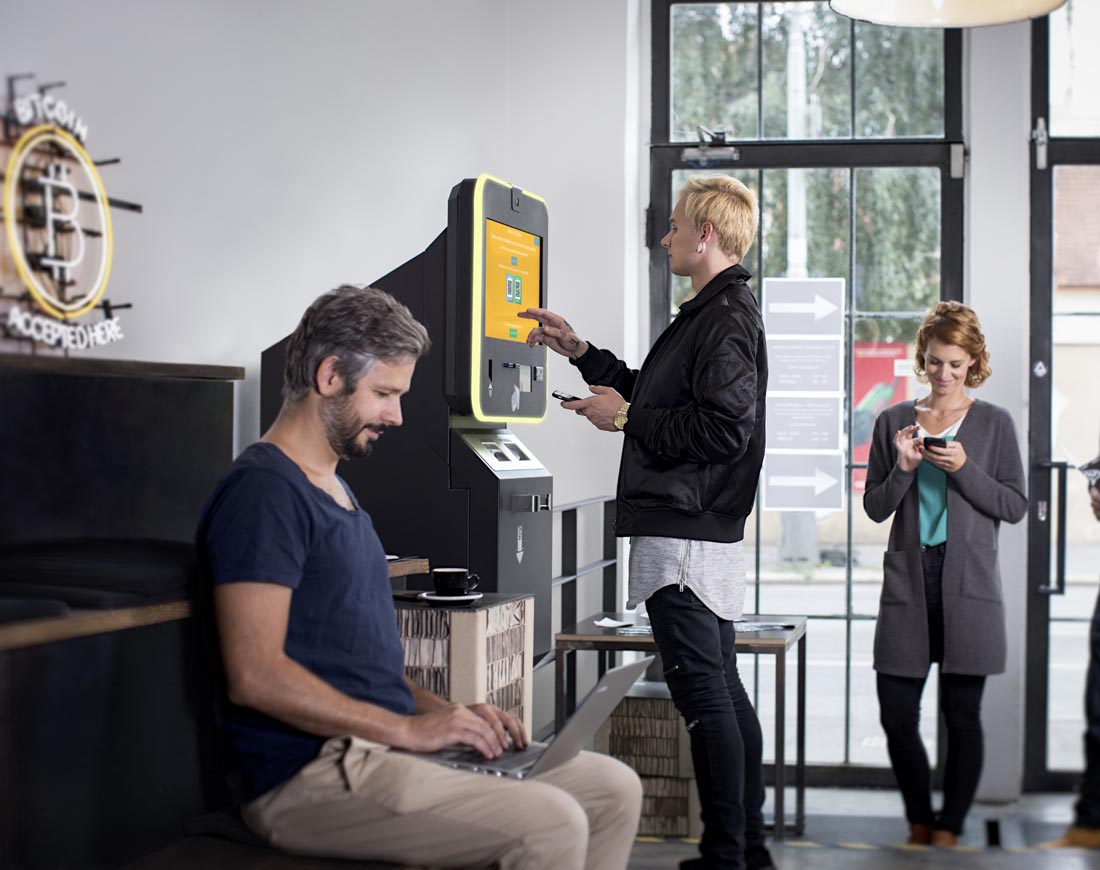 Eine Person steht mit Smartphone an einem Bitcoin-Automaten, hinter ihr warten Leute