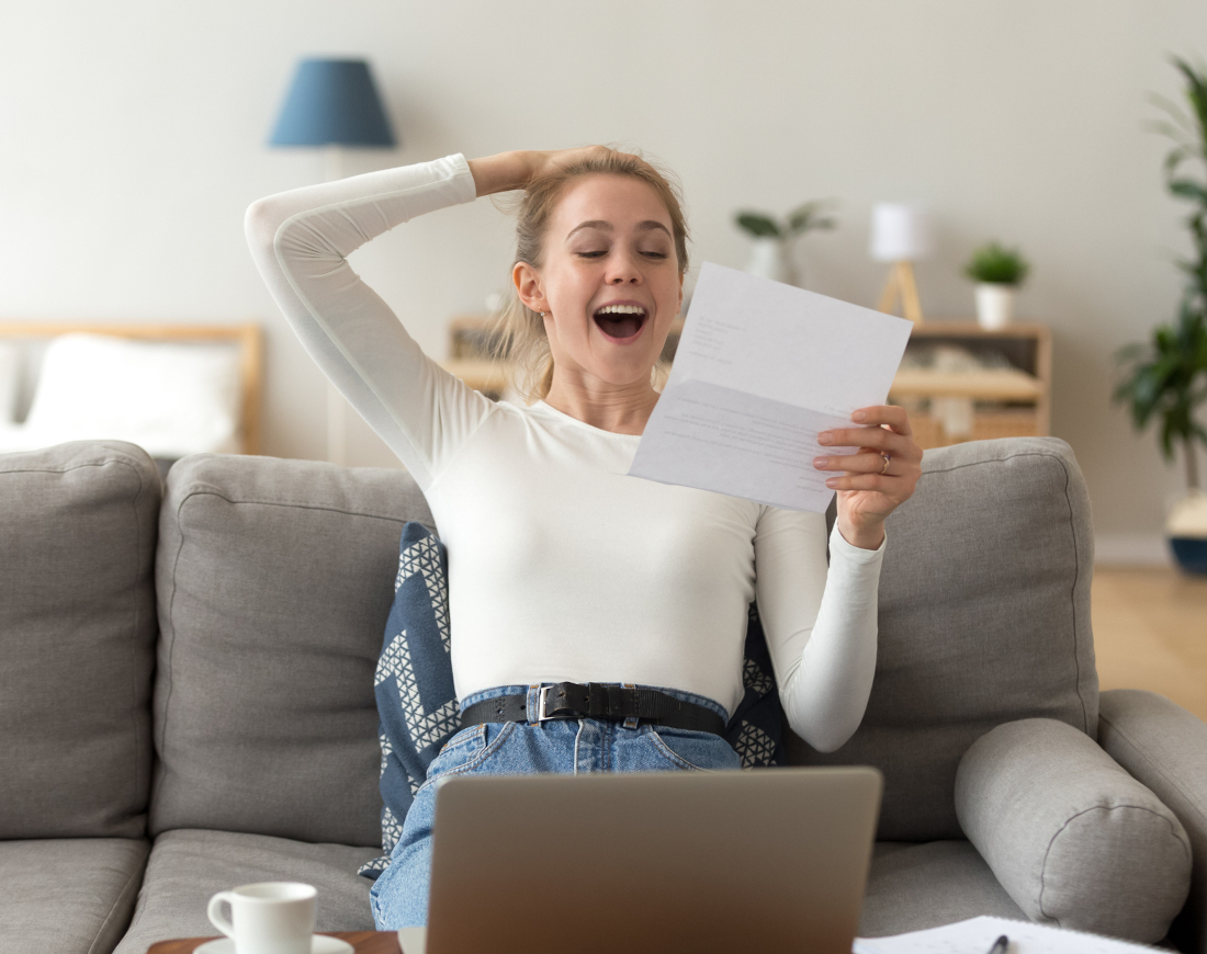 Eine junge Frau sitzt, ein Schreiben über eine Sonderzahlung in der Hand haltend, freudestrahlend auf dem Sofa