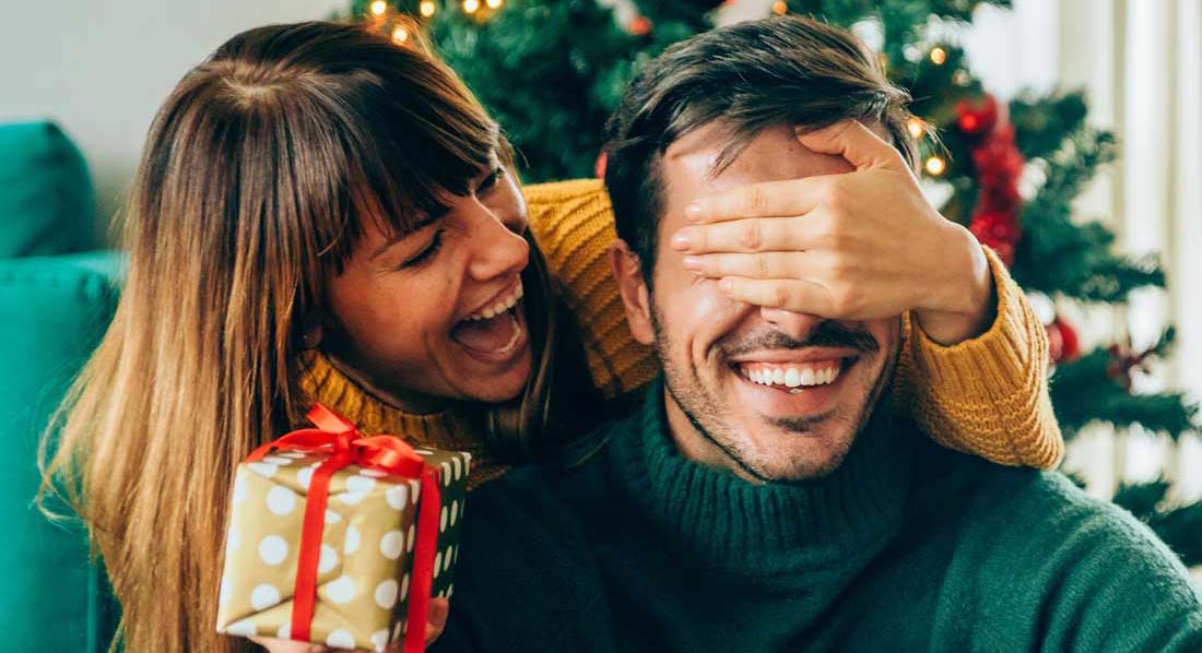 Eine junge Frau hält ihrem Freund von hinten die Augen zu, in der Hand ein Geschenk.