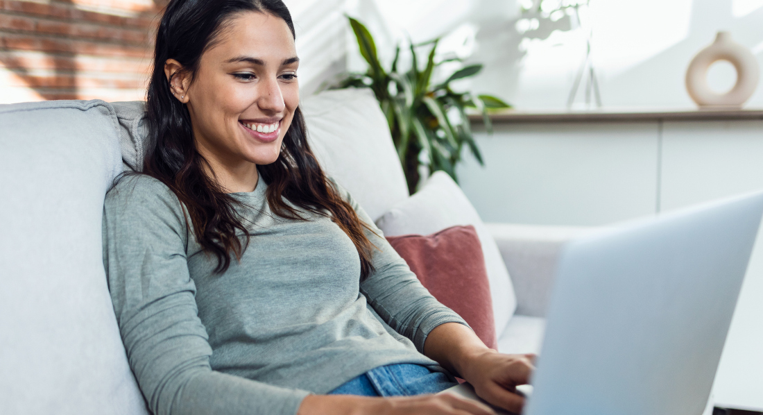 Junge Frau sitzt entspannt auf dem Sofa und tippt auf ihrem Laptop