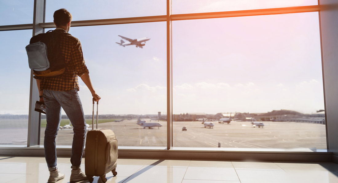 Reisender mit Koffer blickt vom Flughafenterminal auf die Landebahn und beobachtet ein startendes Flugzeug