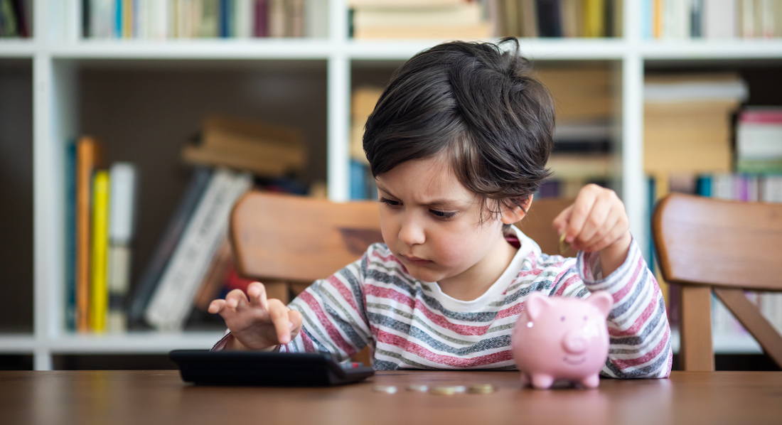Kleines Mädchen wirft mit der einen Hand eine Münze in ein Sparschwein und rechnet die angesparte Summe mit der anderen Hand auf dem Taschenrechner zusammen