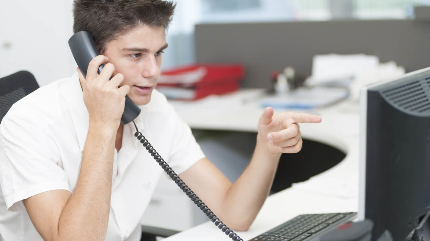 Ein telefonierender Jugendlicher sitzt an einem Büroarbeitsplatz und deutet mit der linken Hand auf den Monitor vor ihm
