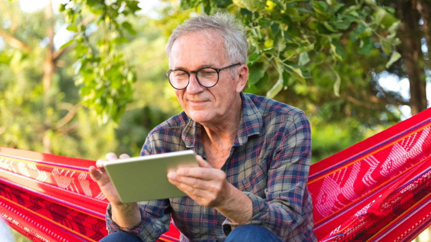 Ein Mann mit grauen Haaren sitzt auf einer Hängematte im Garten mit einem Tablet in der Hand