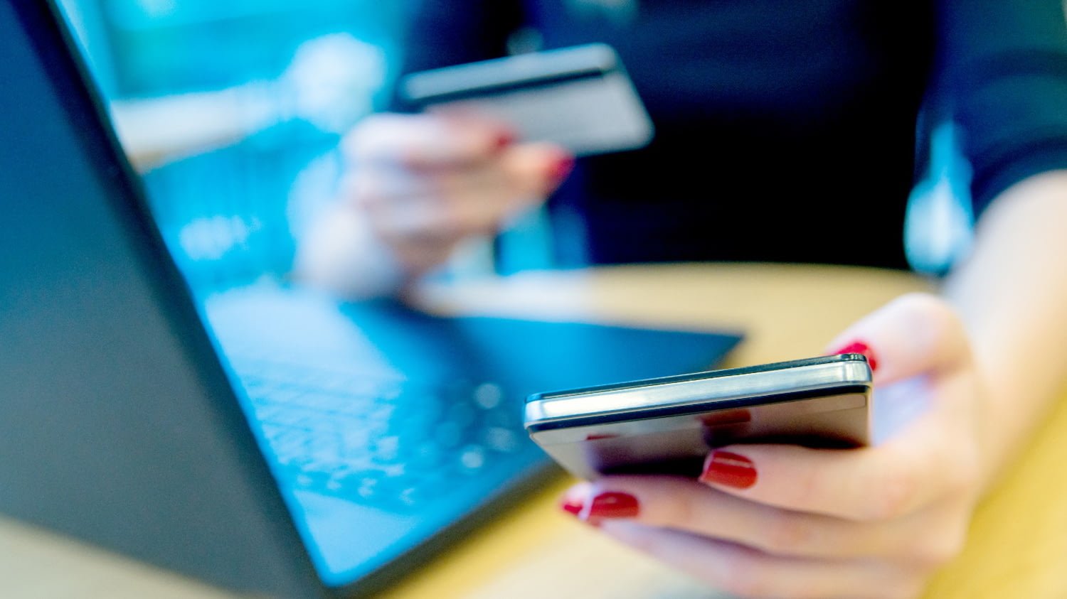 Eine Damenhand tippt in Großaufnahme auf dem Smartphone vor einem Laptop, die andere hält eine Kreditkarte.