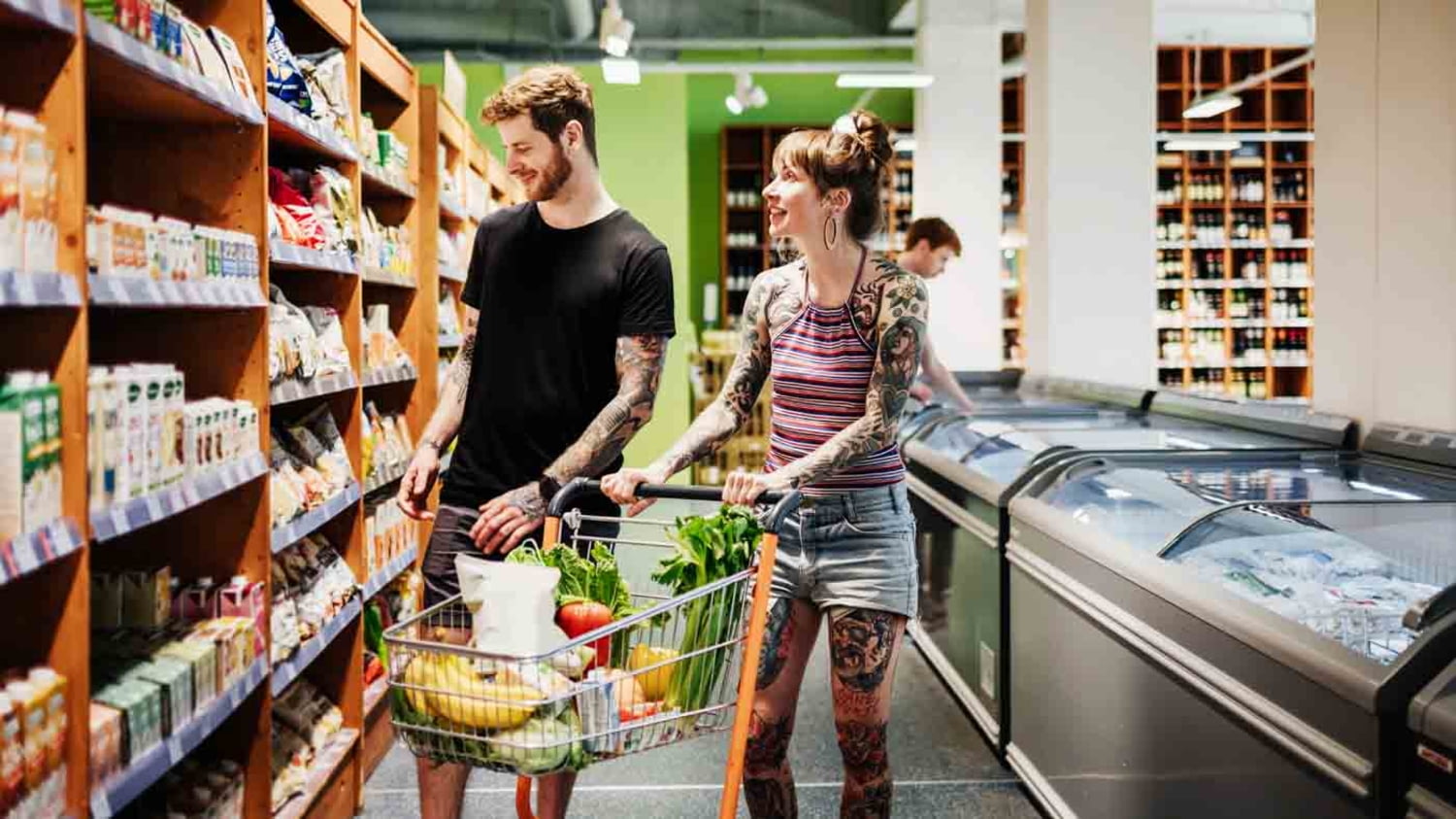 Ein junges, tätowiertes Paar steht mit einem gefüllten Einkaufswagen vor einem Supermarktregal und betrachtet die Auslage