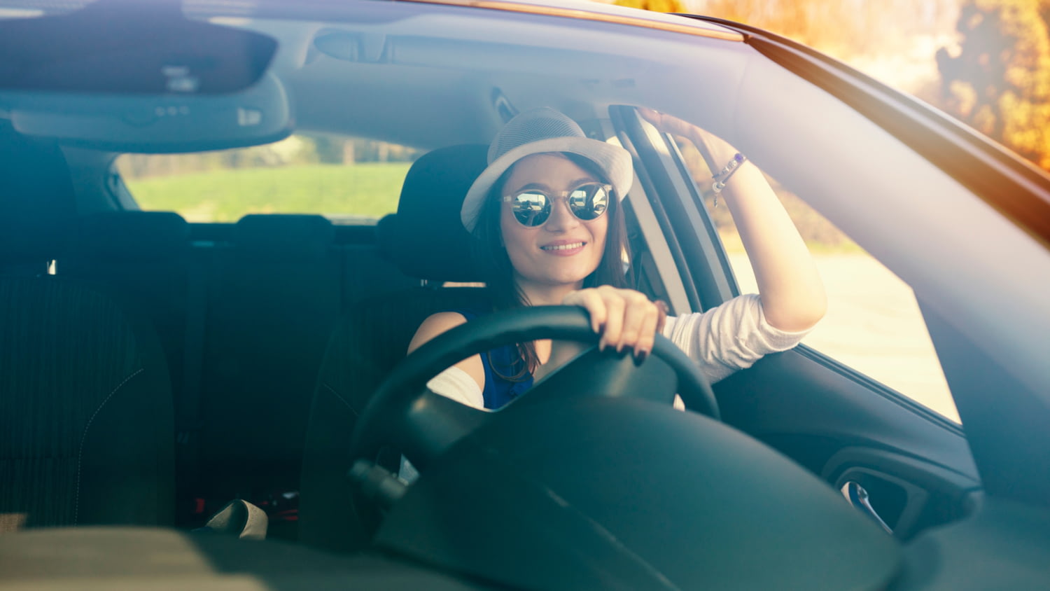 Eine junge Frau mit Sonnenbrille und Hut fährt lächelnd in einem Auto