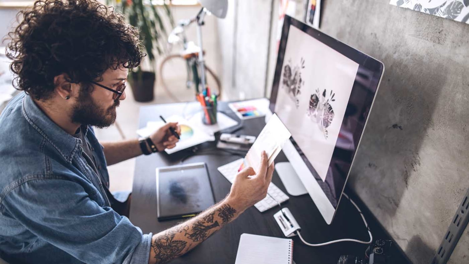 Junger Kreativer an seinem Schreibtisch mit Zeichenstift, Tablet, Monitor und Arbeitsmaterialien