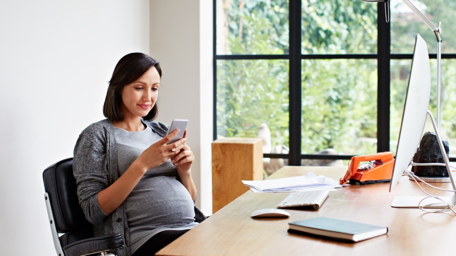 Eine junge schwangere Frau sitzt am Schreibtisch und schaut auf ihr Smartphone