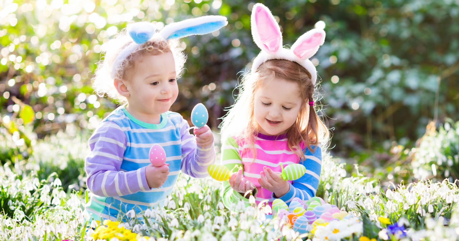 Zwei kleine Kinder mit Hasenohren-Haarreifen suchen Ostereier in einem Blumenfeld
