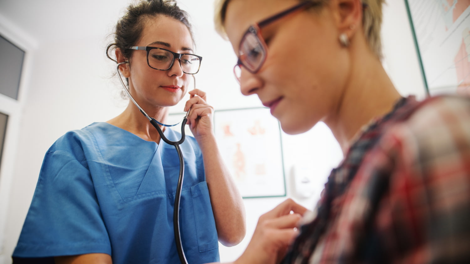 Stehende Frau in blauer OP-Bekleidung hört mit einem Stethoskop die Brust einer sitzenden Frau ab