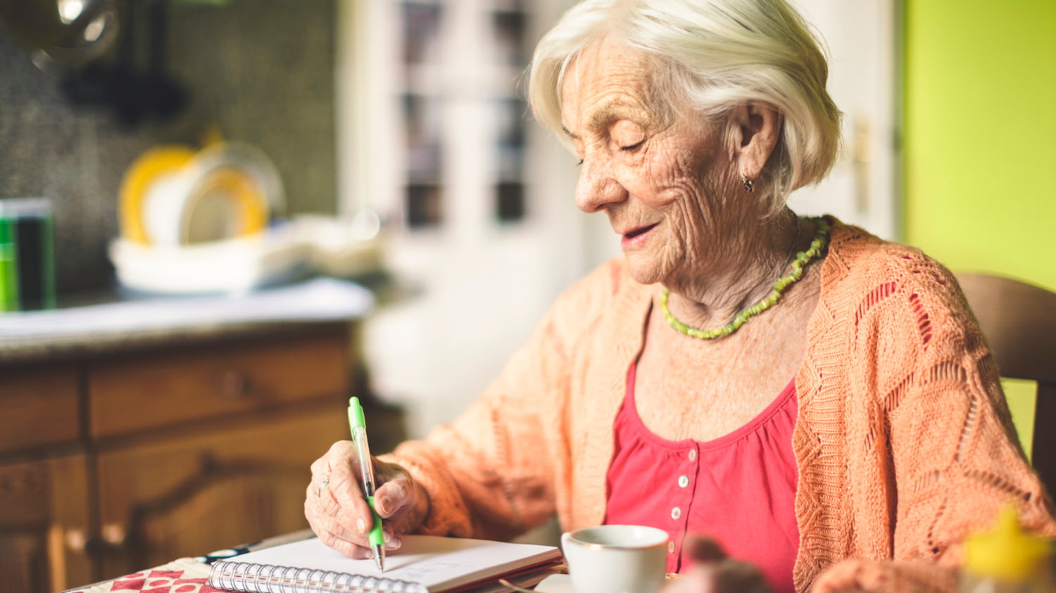 Eine Seniorin sitzt in der Küche und schreibt etwas in einen Spiralheft