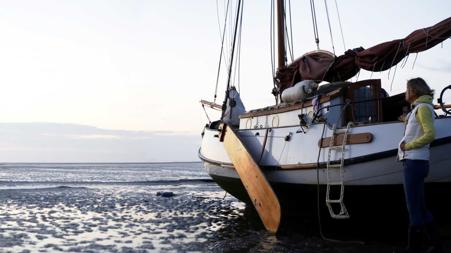 Im Wattenmeer auf Grund gelaufenes Segelboot, daneben eine Frau, die Richtung Horizont schaut