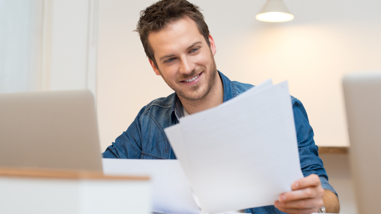 Ein junger Mann sitzt am Schreibtisch vor einem Laptop und schaut sich l&auml;chelnd Dokumente an