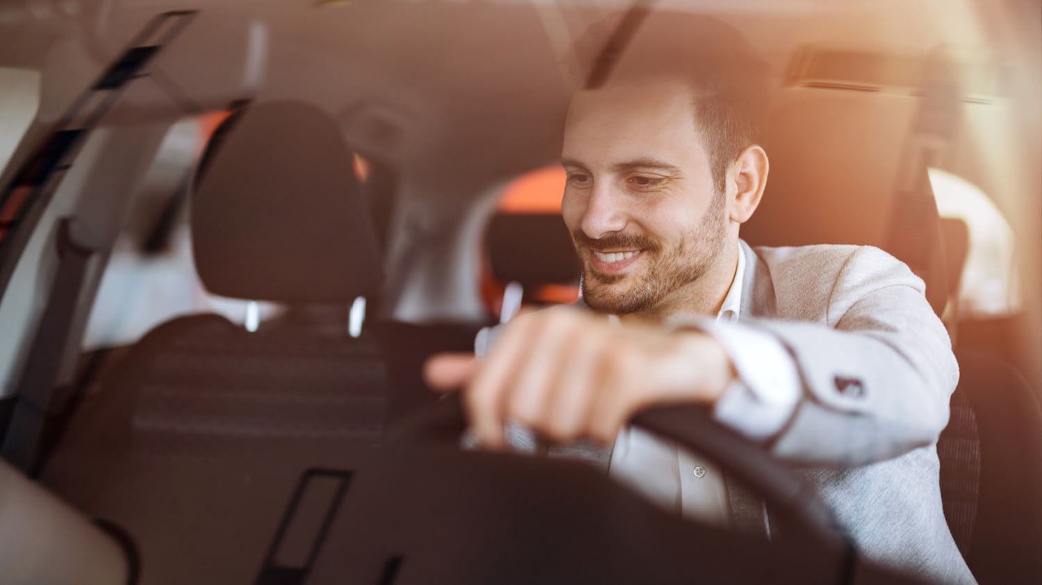 Ein jüngerer Mann im Business-Outfit sitzt gut gelaunt am Steuer eines Autos.