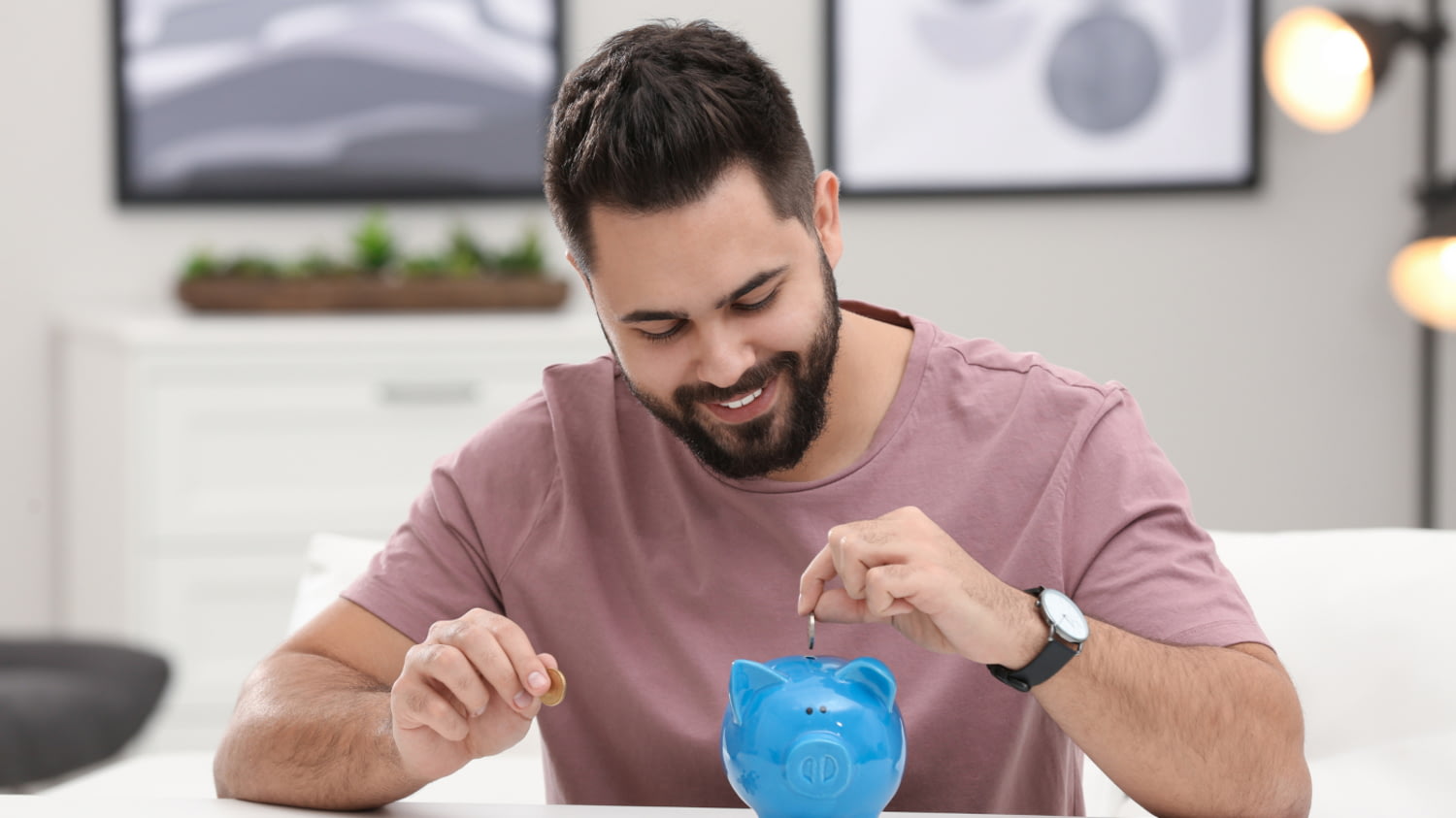 Ein Mann mit Bart sitzt am Tisch und blickt lächelnd auf ein blaues Sparschwein, in das er eine Münze steckt