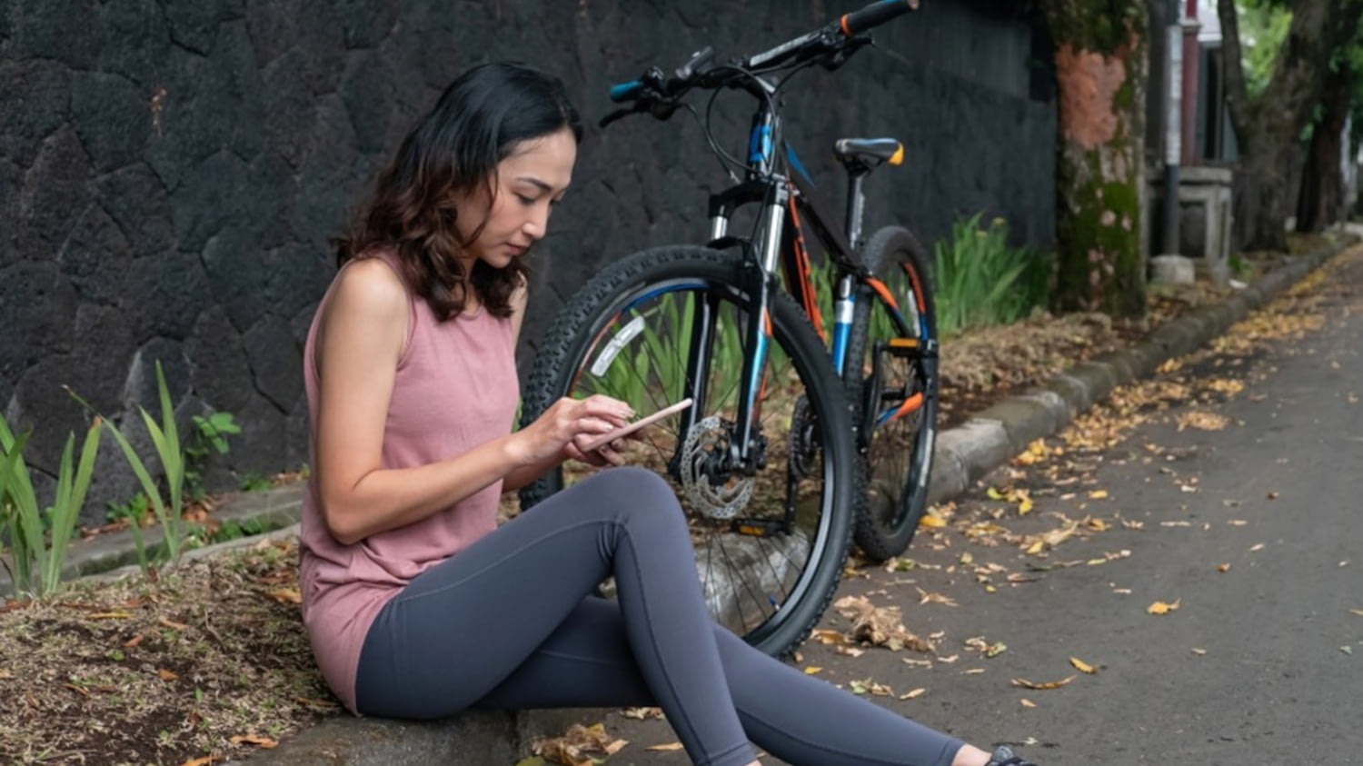 Junge Frau hat eine Fahrradpanne, sitzt auf dem Bürgersteig und gibt etwas auf dem Smartphone ein