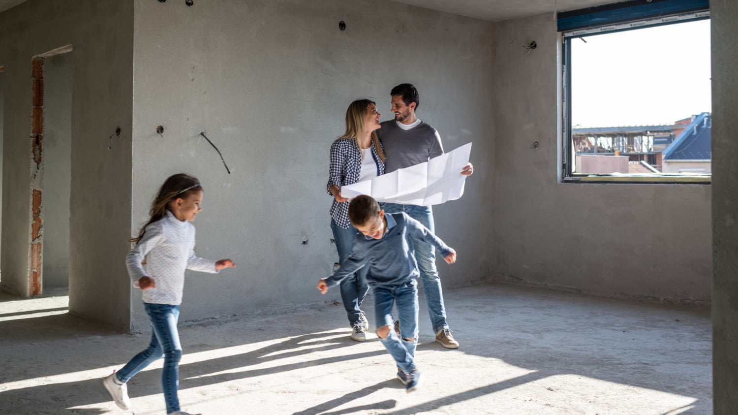 Junge Familie mit zwei Kindern im Rohbau eines Hauses betrachtet einen Bauplan