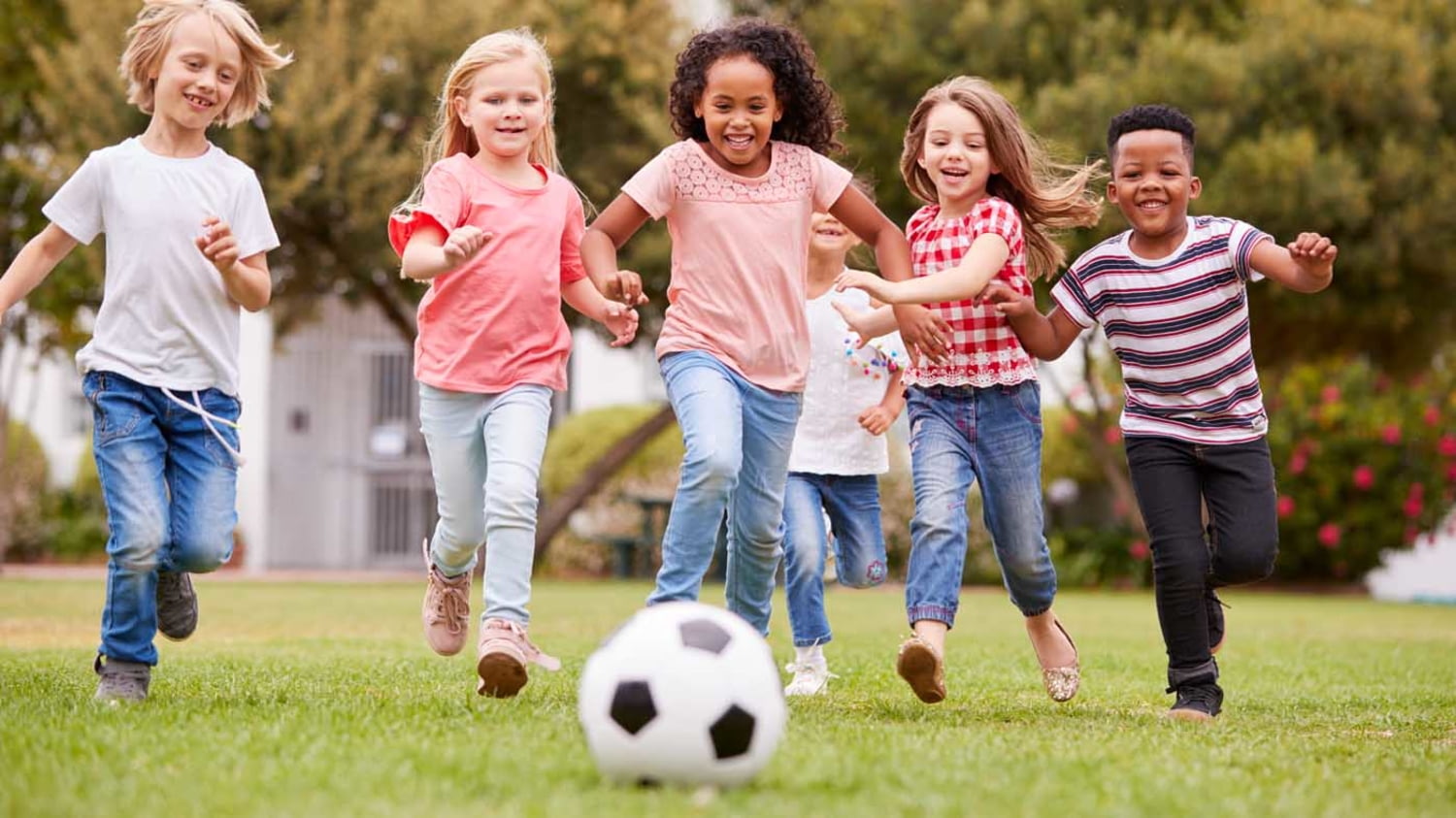 Sechs Kinder laufen lachend auf einen Fußball im Vordergrund zu
