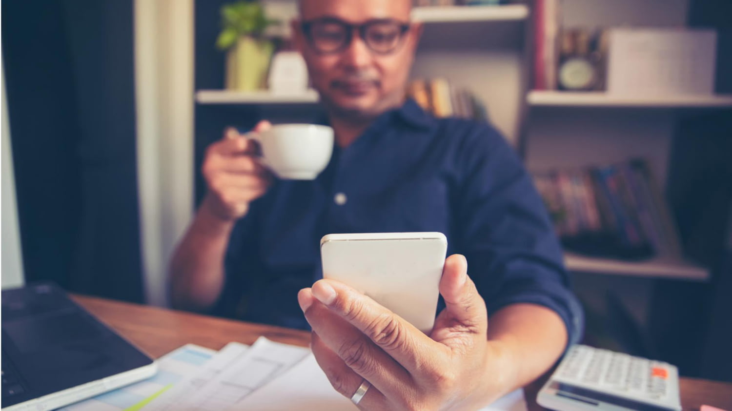 Mann trinkt Kaffee am heimischen Schreibtisch und blickt auf sein Smartphone