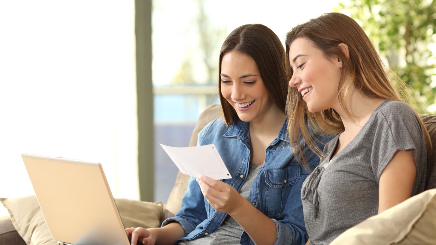 Zwei junge Frauen sitzen auf einem Sofa und schauen lachend auf einen Laptop und einen Kontoauszug.