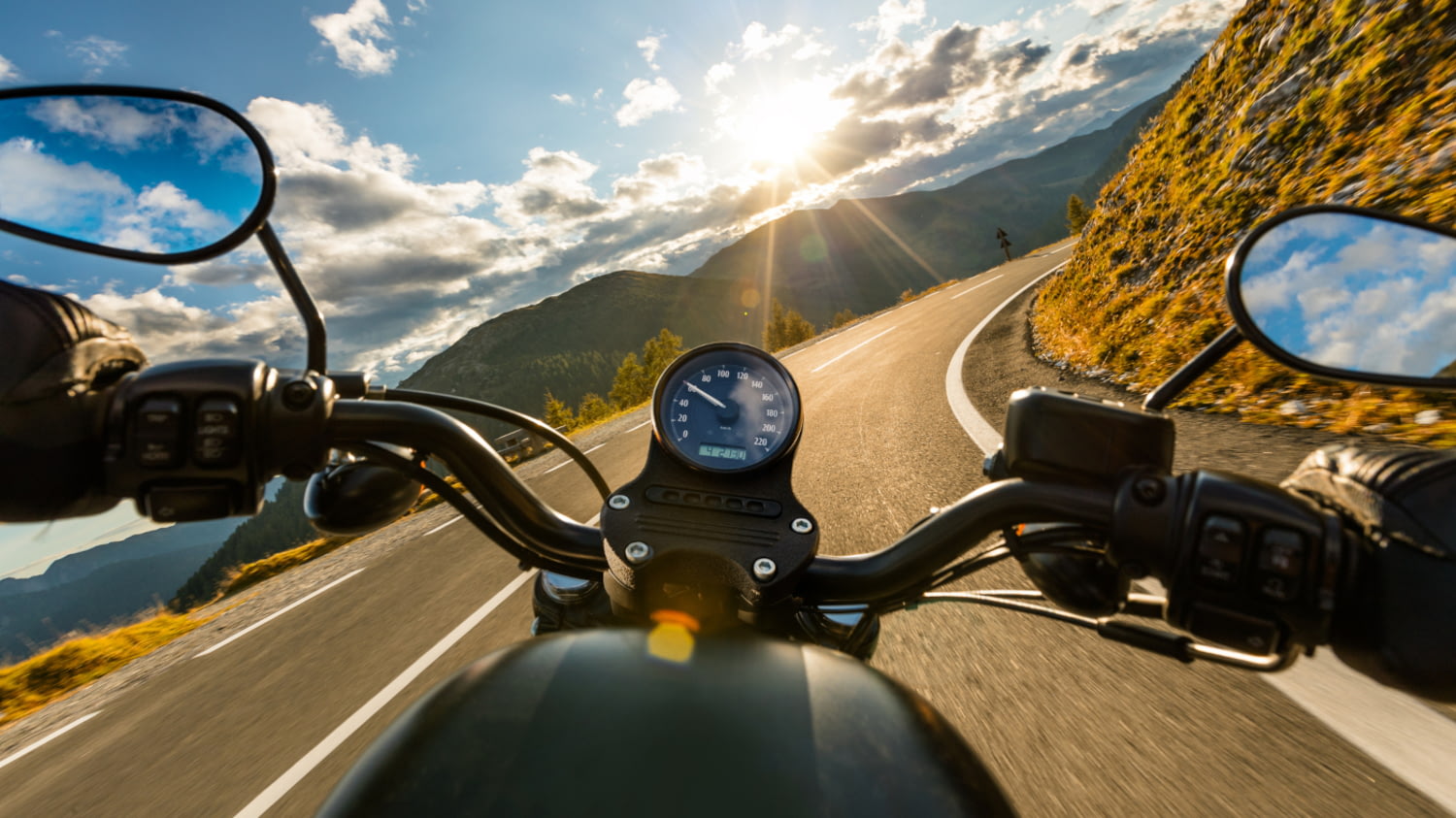 Motorradfahrer*in-Perspektive über einen Lenker auf eine Straße in den Bergen bei sonnigem Wetter