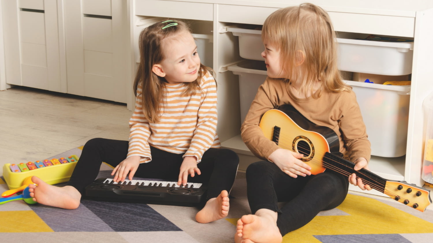Zwei kleine Mädchen sitzen barfuß auf dem Boden, spielen mit Keyboard und Gitarre
