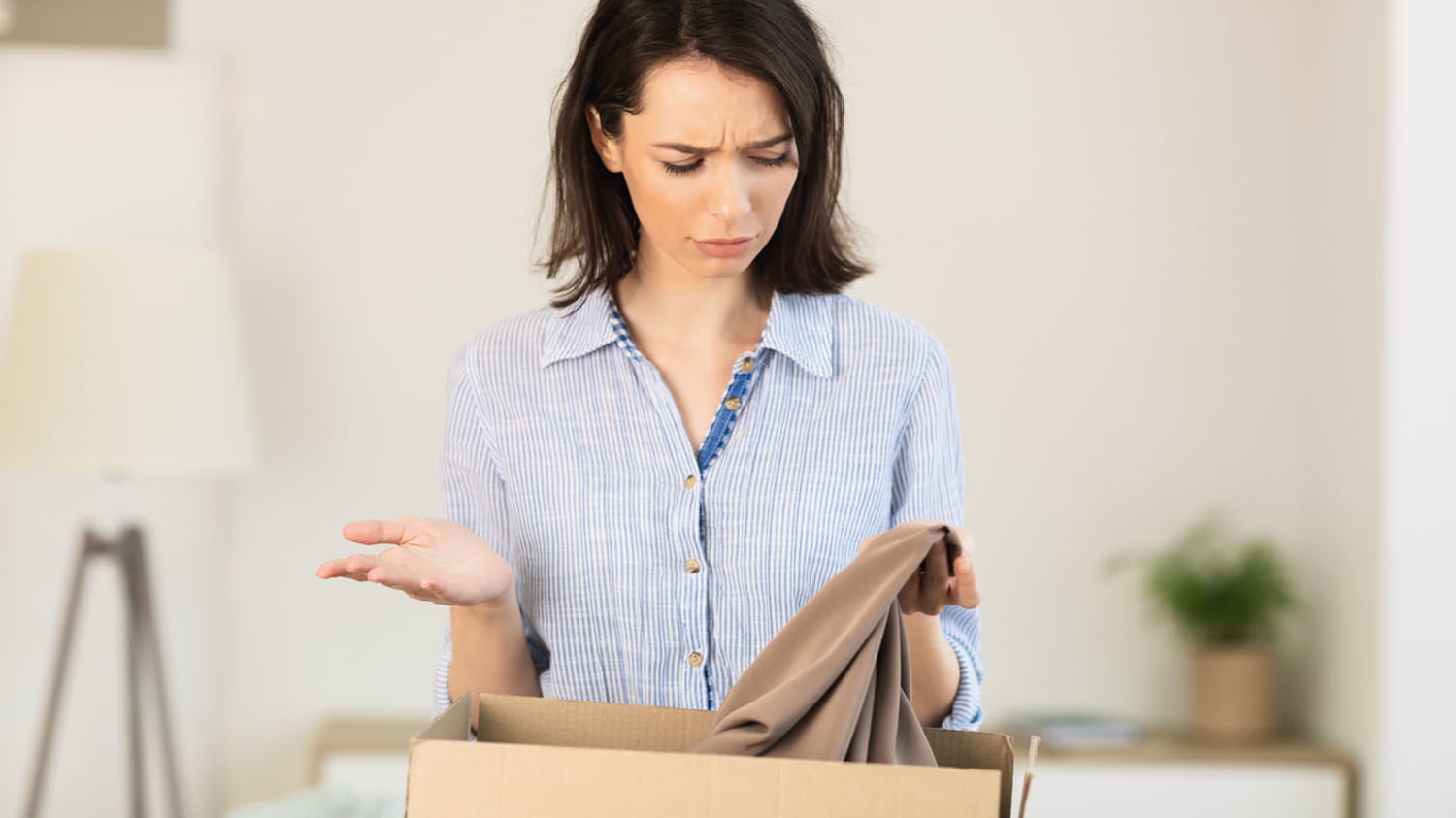 Junge Frau betrachtet mit kritischem Blick ein Kleidungsstück, das sie aus einem Paket ausgepackt hat