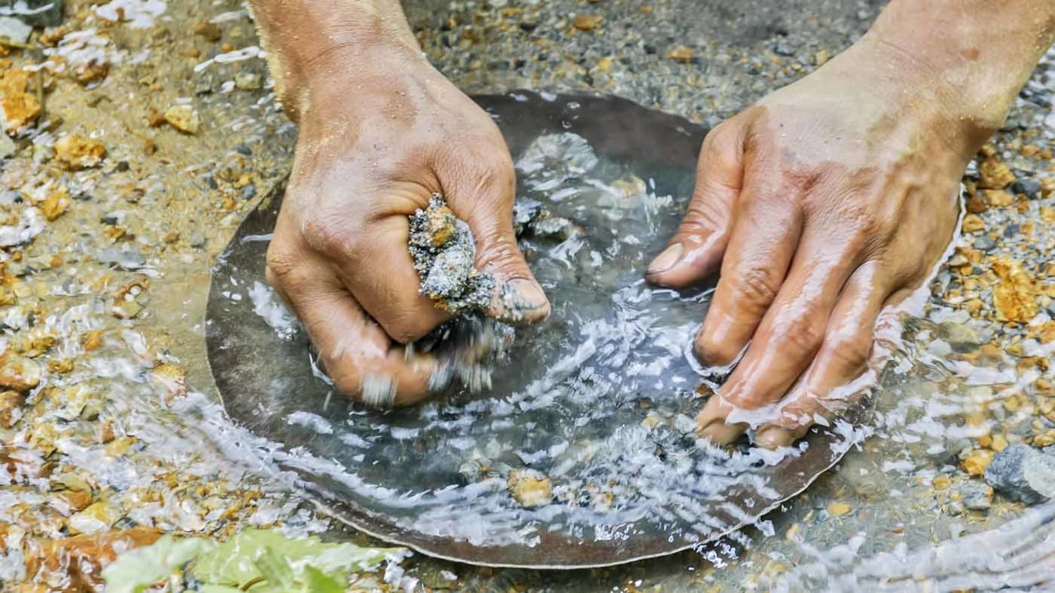 Nahaufnahme zweier Hände, die mithilfe einer Goldwaschpfanne in einem Flussbett nach Gold suchen