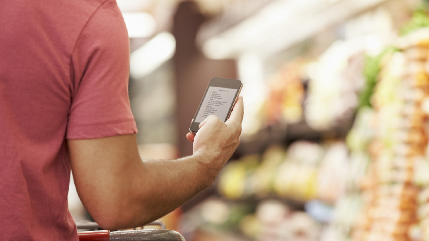 Ein Mann mit Einkaufswagen im Supermarkt hält in der rechten Hand ein Smartphone, auf dem eine Liste zu sehen ist