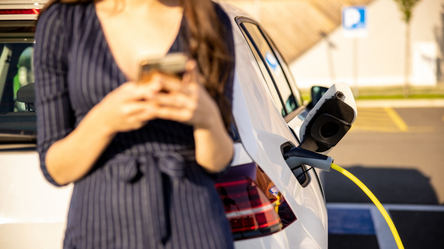 Junge Frau mit Handy in der Hand, während sie auf das Aufladen des Elektroautos wartet