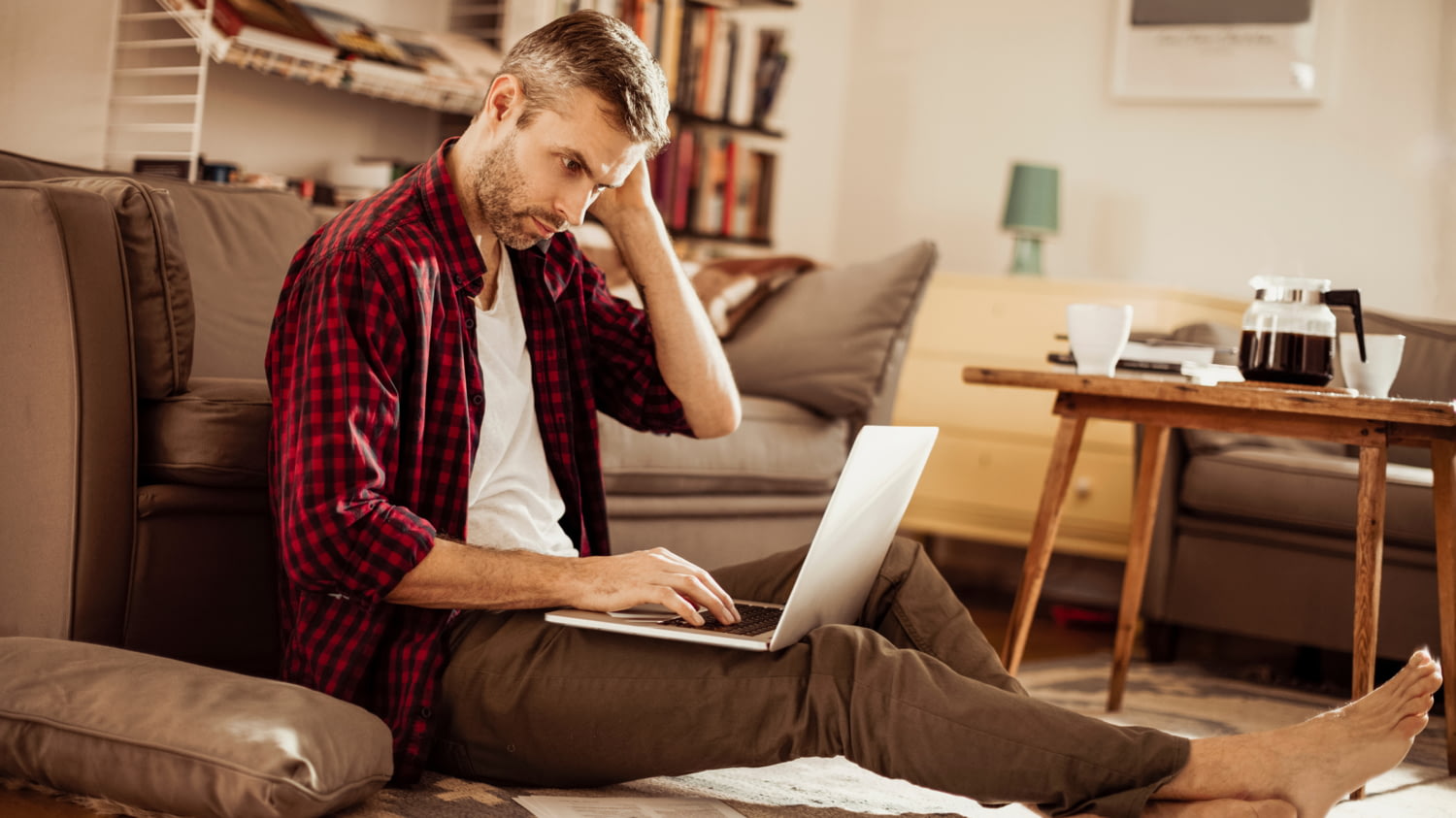 Ein Mann mit Laptop auf dem Schoß sitzt mit ausgestreckten Beinen auf dem Boden eines Wohnzimmers