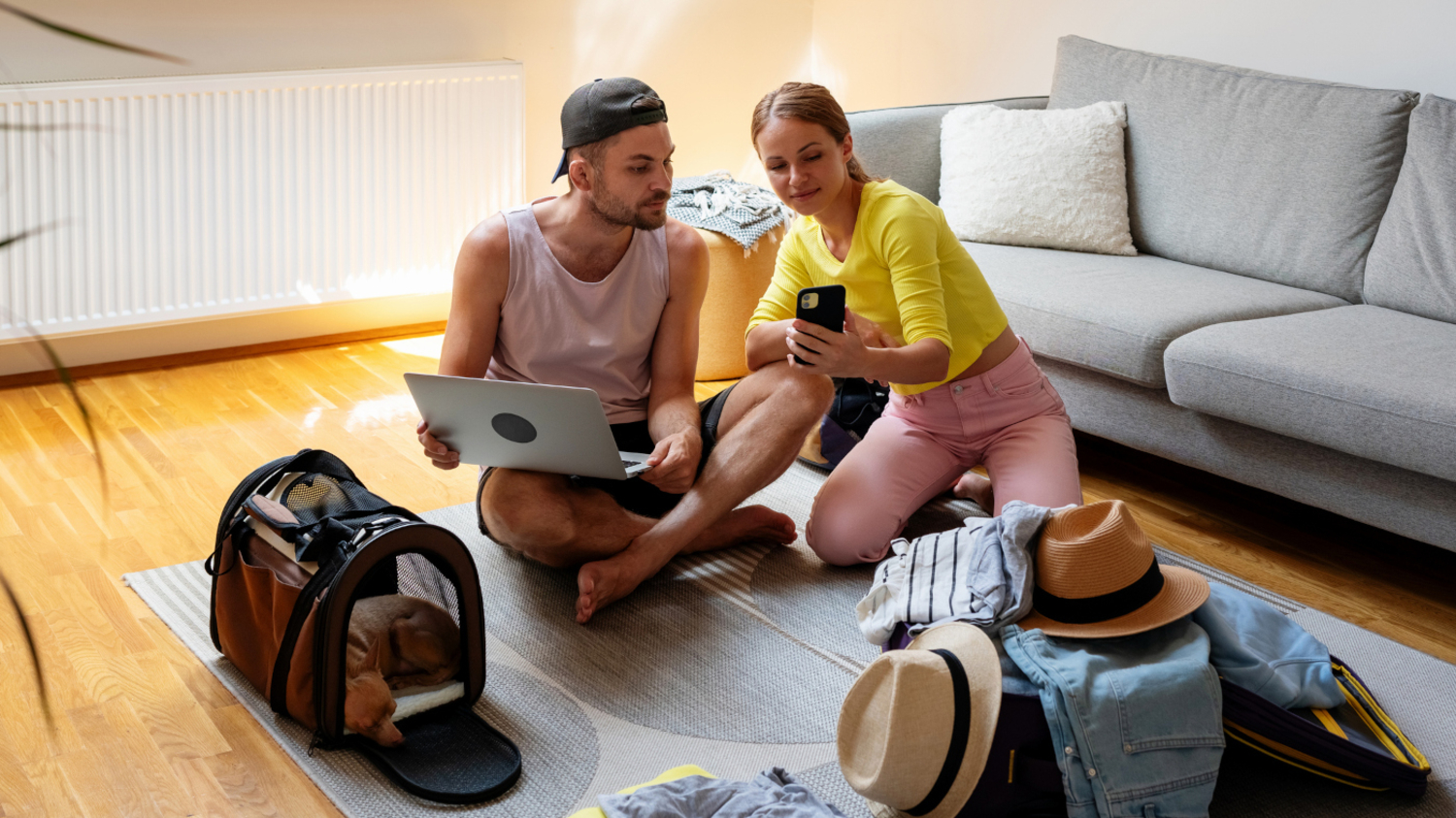 Ein junges Paar sitzt im Wohnzimmer und sucht auf dem Handy und dem Laptop nach Urlaubsreisen mit Frühbucherrabatt