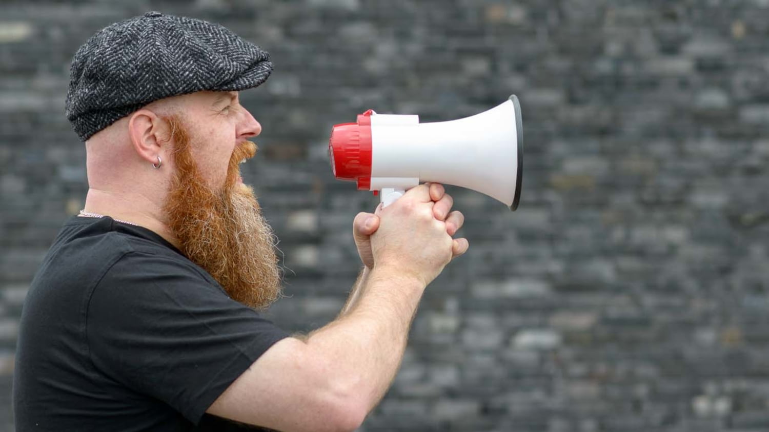 Bärtiger Mann steht vor einer Wand und schreit in ein Megafon