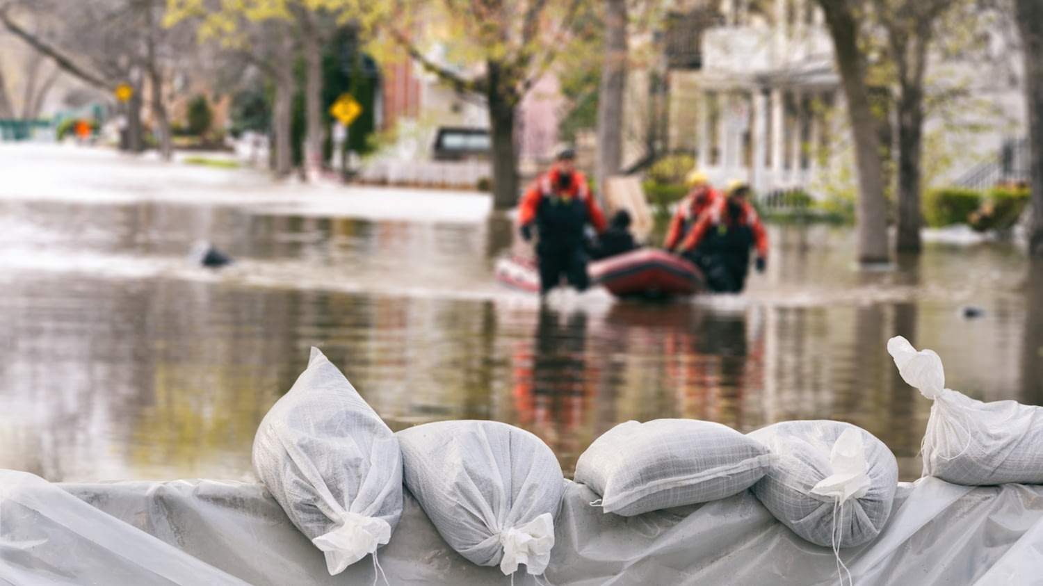 Sandsäcke im Vordergrund sollen Häuser schützen, dahinter ein Schlauchboot mit Helfern auf einer überschwemmten Straße