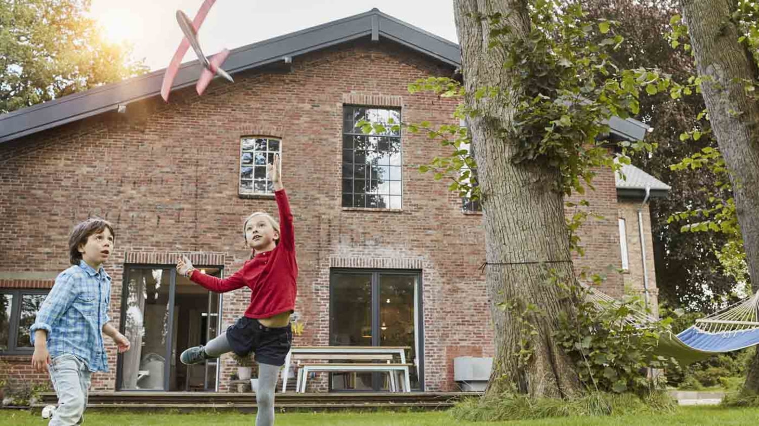 zwei Kinder spielen im Garten vor einem Backsteinhaus mit einem Modellflugzeug