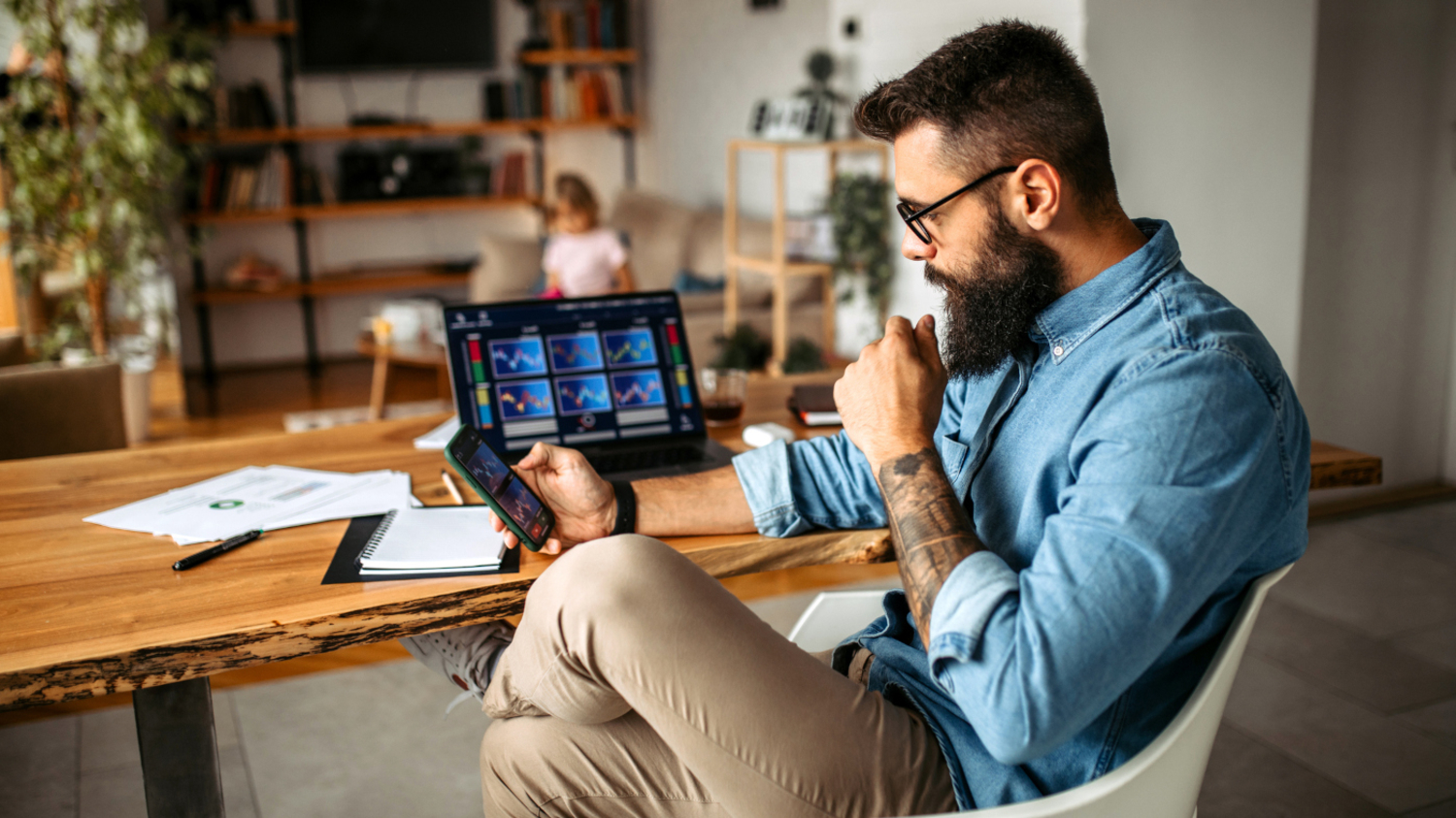 Ein Mann sitzt an seinem Wohnzimmertisch und beobachtet B&ouml;rsenkurse auf dem Smartphone und auf dem Laptop