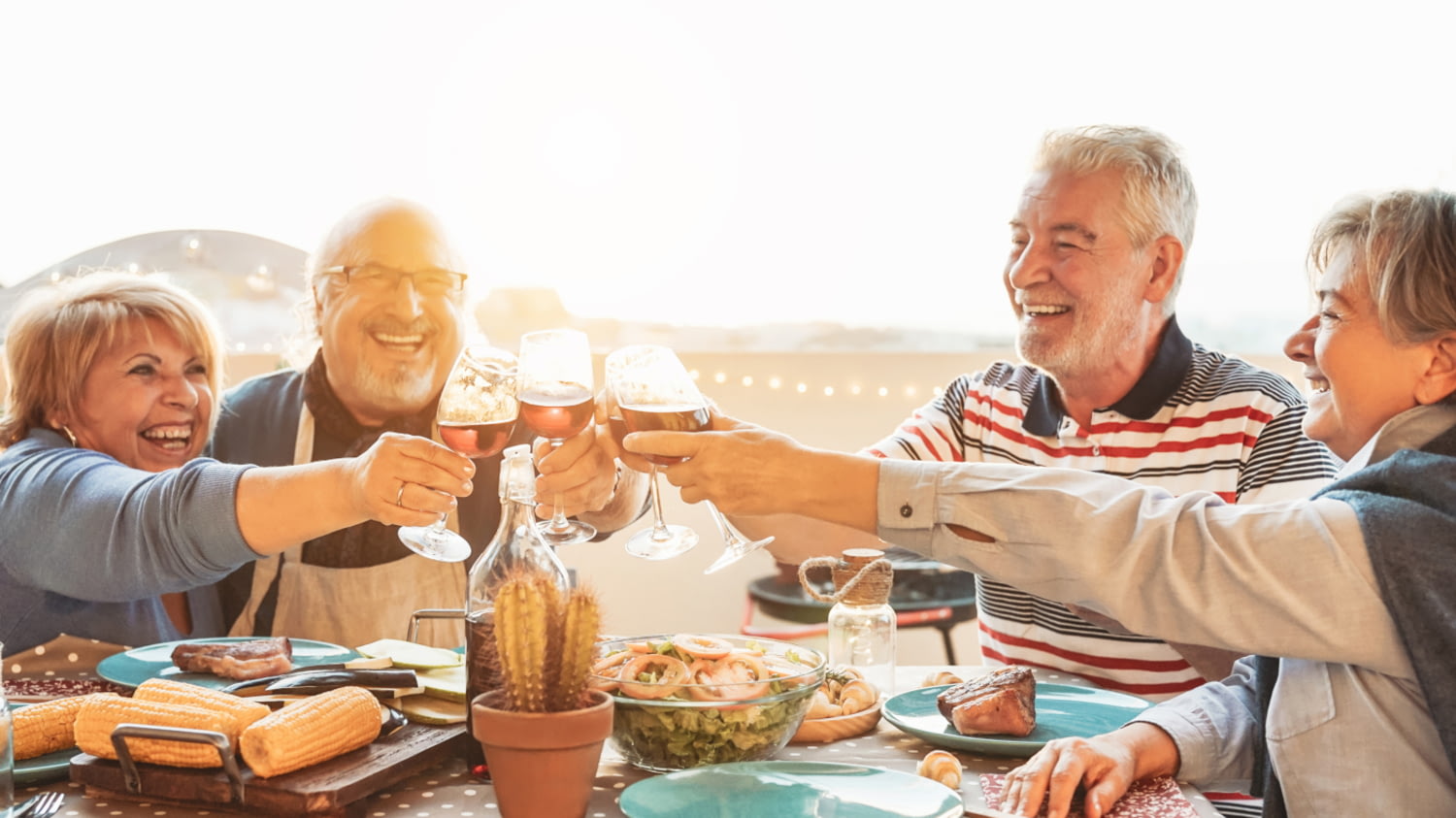 Glückliche Rentner*innen stoßen mit Rotweingläsern bei einem Barbecue-Dinner an