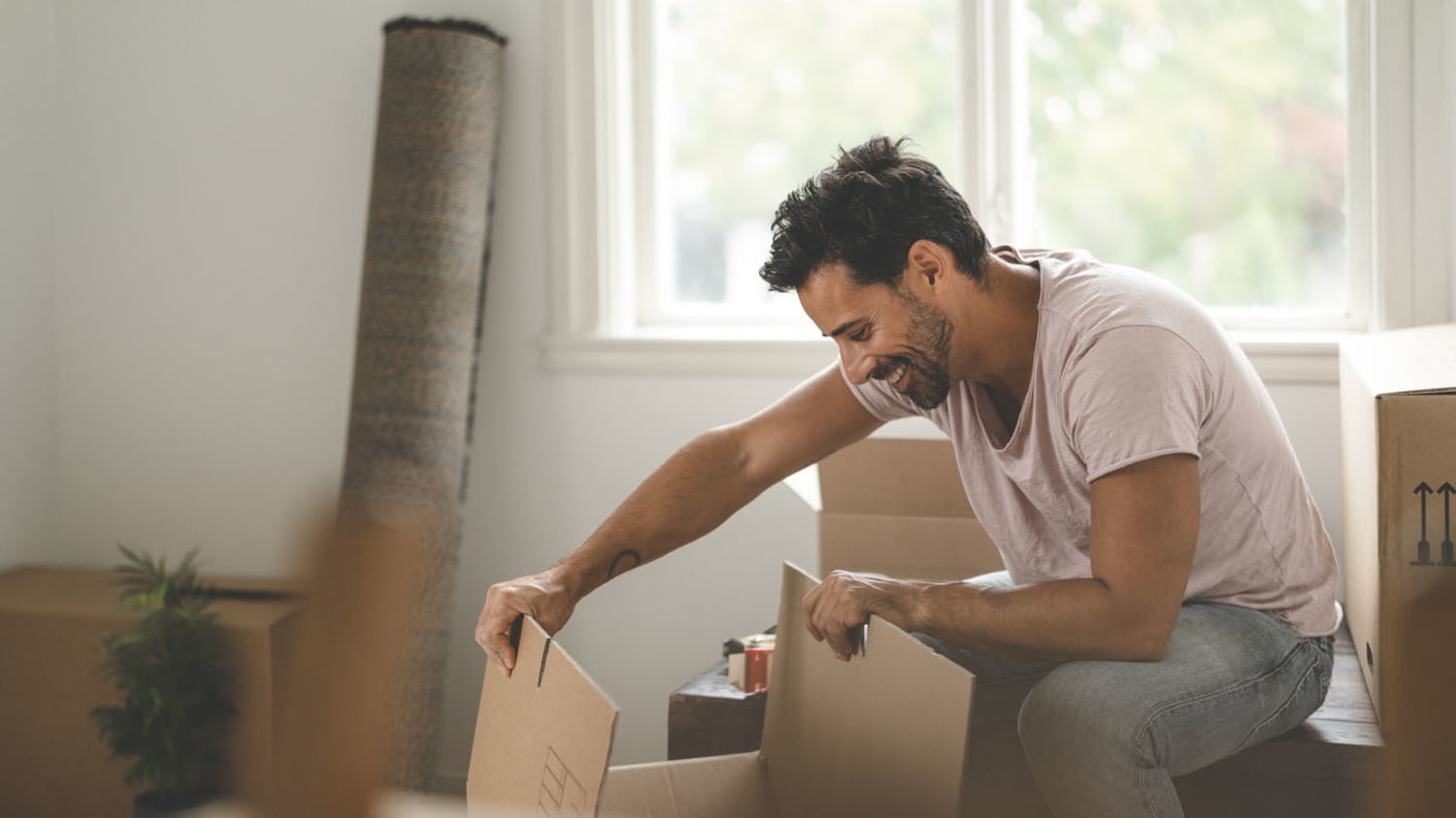 Ein Mann in Jeans und T-Shirt schaut in seiner neuen Wohnung lächelnd in einen Umzugskarton