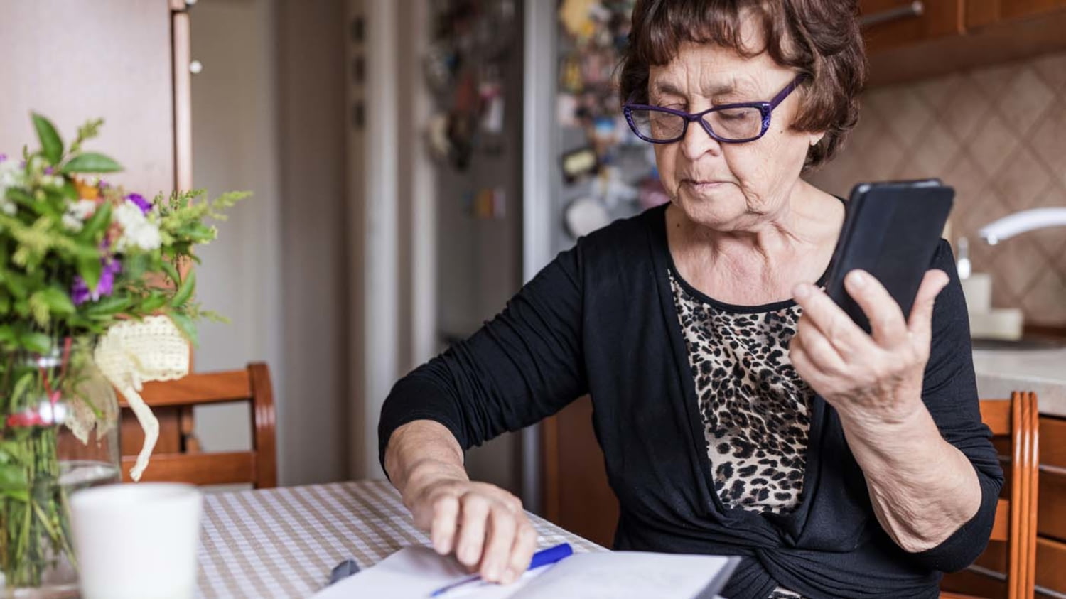 Eine ältere Frau sitzt am Küchentisch und schaut auf ein Notizbuch, während sie ihr Handy in der Hand hält