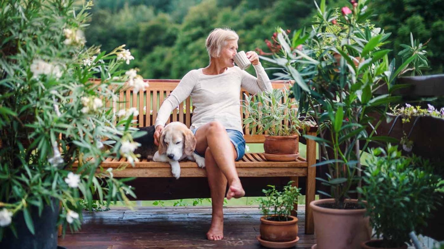 Seniorin sitzt auf der Terrasse auf einer Gartenbank und trinkt einen Kaffee, neben ihr auf der Bank sitzt ein Hund