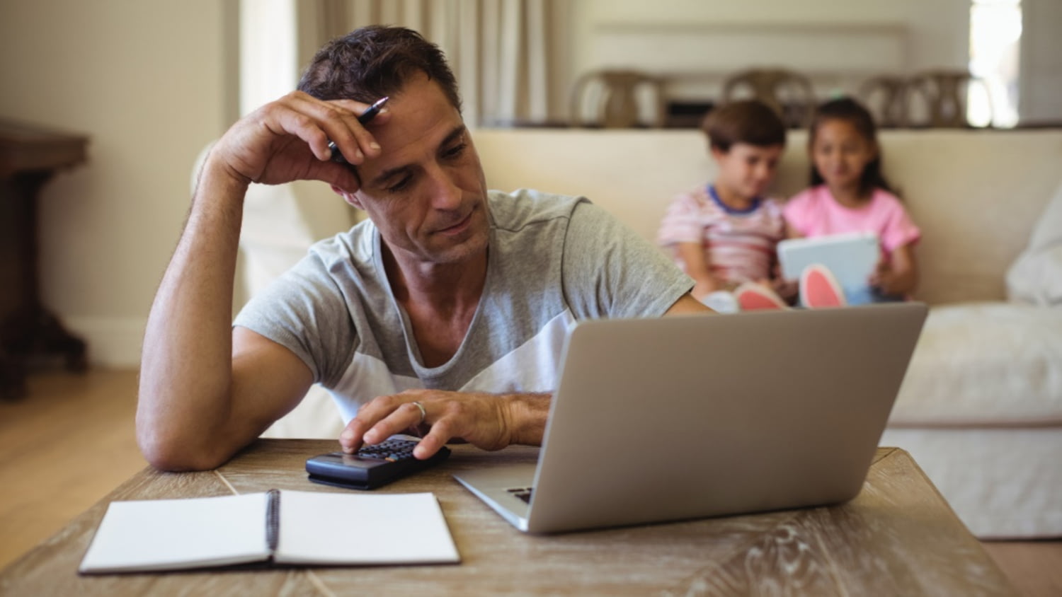 Ein Mann sitzt am Tisch und berechnet sein Haushaltseinkommen, im Hintergrund seine zwei Kinder