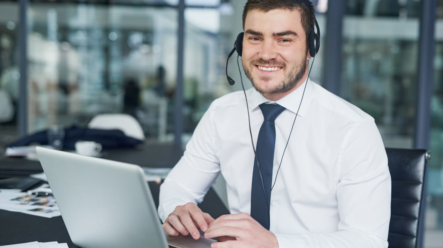 Ein lächelnder Kundendienstberater sitzt mit Laptop und Headset am Schreibtisch