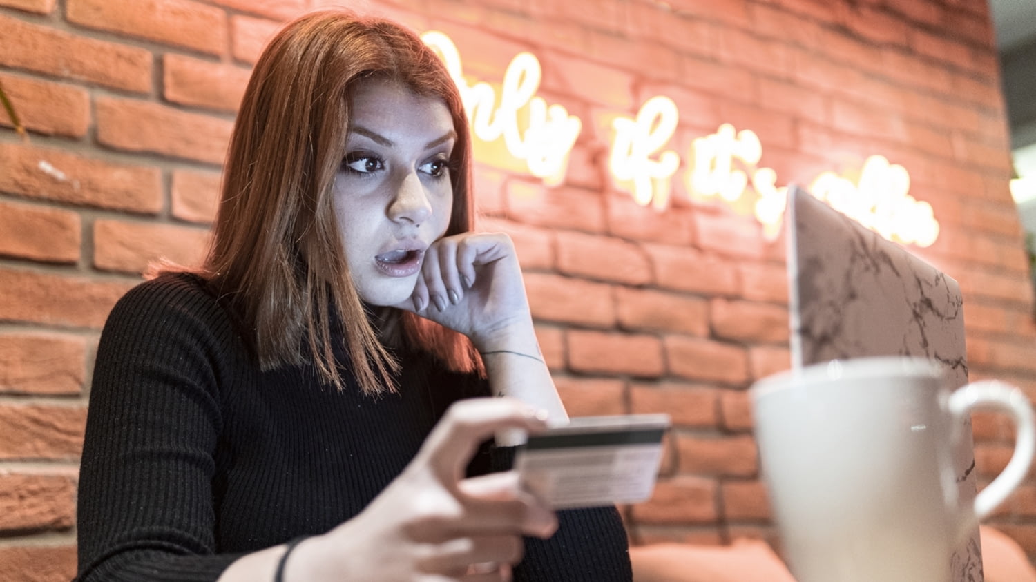 Junge Frau in einem Café blickt erstaunt auf die Kreditkartenabrechnung auf ihrem Laptop