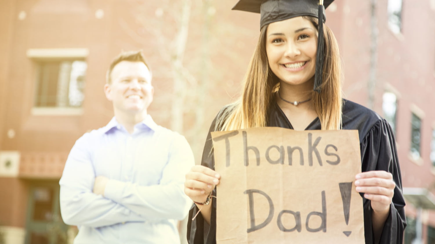 Studentin hält bei ihrer Abschlussfeier ein Schild mit der Aufschrift „Thanks Dad“, im Hintergrund der stolze Vater