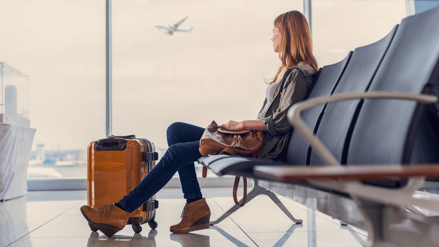 Junge Frau mit Rollkoffer schaut aus der Abflughalle einem startenden Flugzeug hinterher.