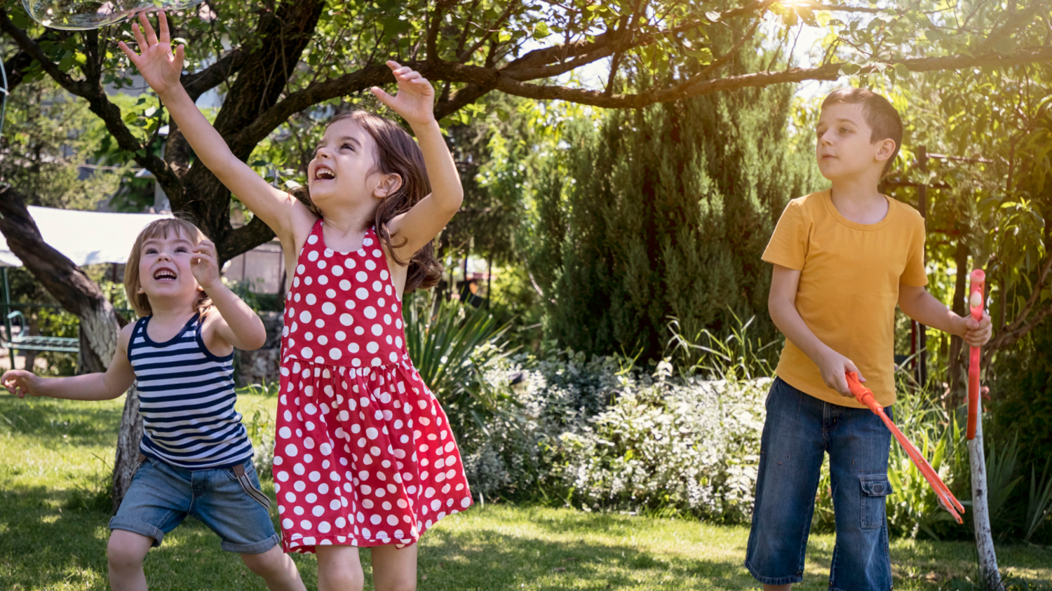 Drei Kinder spielen im Sommergarten und laufen riesigen Seifenblasen hinterher